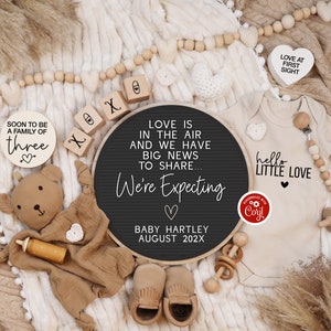 K&ouml;nnte beinhalten: Ein schwarzes Buchstabenbrett mit dem Text "Love is in the air and we have big news to share... We're Expecting" mit einem Herz und dem Text "Baby Hartley August 202X". Es gibt Holzbl&ouml;cke, die "XOXO" buchstabieren, und ein Holzschild, auf dem "Soon to be a family of three" mit einem Herz steht. Es gibt einen Teddyb&auml;r, einen Baby-Strampler mit dem Text "hello little love" und einem Herz und eine Holzrassel.