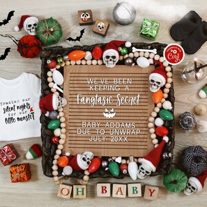 Op de afbeelding: Een baby-aankondiging met Halloween-thema met een houten bord waarop staat "We've been keeping a fangtastic secret. Baby Addams due to unwrap July 20XX". Het bord is omringd door verschillende Halloween-decoraties, waaronder schedels, pompoenen en lampjes.