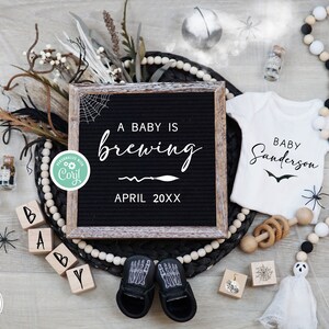May include: A Halloween-themed baby announcement with a black letter board that reads "A baby is brewing April 20XX". There are black and white baby booties, a white onesie with the text "Baby Sanderson", and other Halloween decorations.