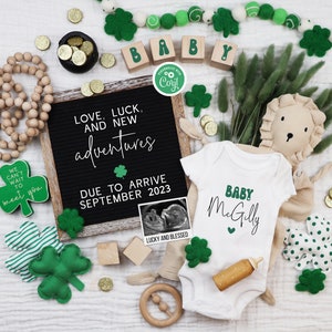 May include: A white onesie with the text "Baby McGilly" in green and a small heart. The onesie is surrounded by green shamrocks, a wooden lion, and a wooden rattle. A black letter board with the text "Love, Luck, and New Adventures Due to Arrive September 2023" is in the background.