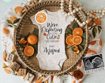 May include: A white babygrow with the text "We're Expecting a Little Cutie This February Baby Ashford" and a small orange graphic. The babygrow is surrounded by orange slices, wooden blocks spelling "BABY", and a small ultrasound image with the text "Our Little Cutie".