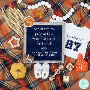 May include: A blue and white letter board with the text "GET READY TO fall in love WITH OUR LITTLE draft pick JOINING THE TEAM NOVEMBER 2022".  A white onesie with the text "Gronkowski 87" is next to the letter board.  A small pumpkin, a wooden star, a wooden rattle, and a pair of white baby booties are on a plaid blanket.