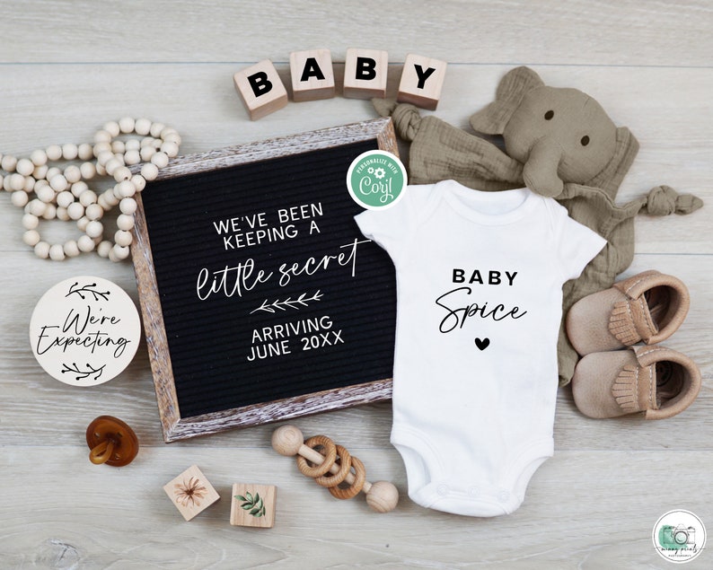 May include: A black letter board with the text "We've been keeping a little secret arriving June 20XX". A wooden round sign with the text "We're Expecting" is on the left. A white onesie with the text "Baby Spice" is in the center. A stuffed elephant toy is on the right.