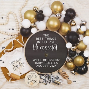 Pode incluir: Um quadro de letras preto com o texto "The best things in life are unexpected" e "We'll be poppin' baby bottles August 202X". O quadro &eacute; rodeado por bal&otilde;es dourados, pretos e brancos. H&aacute; um macac&atilde;o branco com o texto "Countdown to baby Jacobs" e uma placa de madeira com o texto "Soon to be a family of three".