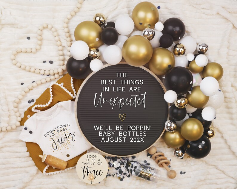 Pode incluir: Um quadro de letras preto com o texto "The best things in life are unexpected" e "We'll be poppin' baby bottles August 202X". O quadro &eacute; rodeado por bal&otilde;es dourados, pretos e brancos. H&aacute; um macac&atilde;o branco com o texto "Countdown to baby Jacobs" e uma placa de madeira com o texto "Soon to be a family of three".