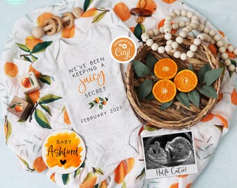 May include: A white onesie with the text "We've been keeping a juicy secret February 2023" and a small orange blossom design. A round orange and white sign says "Baby Ashford" with a small heart. A white and orange blanket with a pattern of oranges and leaves. A small wooden frame with a black and white ultrasound image and the text "Hello Cutie!"