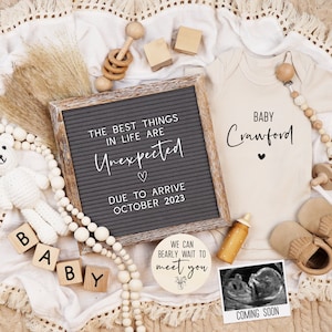May include: A letter board with the text "The best things in life are Unexpected" and "Due to arrive October 2023". A white onesie with the text "Baby Crawford" and a heart. A wooden sign with the text "We can barely wait to meet you". A small white teddy bear, wooden blocks spelling "BABY", a wooden rattle, and a wooden bead garland.