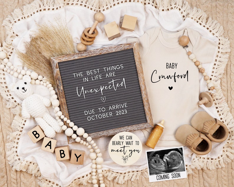 May include: A letter board with the text "The best things in life are Unexpected" and "Due to arrive October 2023". A white onesie with the text "Baby Crawford" and a heart. A wooden sign with the text "We can barely wait to meet you". A small white teddy bear, wooden blocks spelling "BABY", a wooden rattle, and a wooden bead garland.