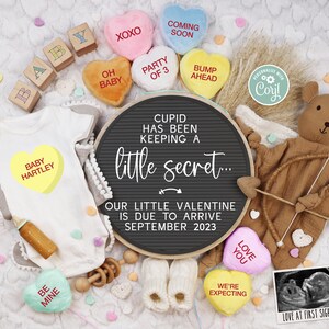 Könnte beinhalten: Eine Schwangerschaftsanzeige zum Valentinstag mit einem Schild aus Schiefer, auf dem steht: "Cupido hat ein kleines Geheimnis gehütet... Unser kleiner Valentinstag wird im September 2023 erwartet.". Das Schild ist umgeben von Babykleidung, einem Teddybär und Herzchen aus Süßigkeiten.