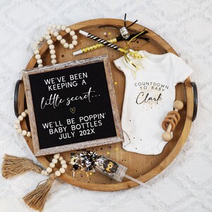 May include: A wooden tray holds a letter board, baby onesie, confetti popper, and wooden beads. The letter board reads "We've been keeping a little secret... We'll be poppin' baby bottles July 202X." The onesie says "Countdown to Baby Clark."