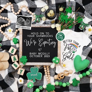 May include: A black and white checkered blanket with a green shamrock and a white onesie with the text "Better Than A Pot Of Gold". There are wooden beads, a wooden sign with the text "Hold On To Your Shamrocks We're Expecting Baby McGilly October 2023", and a small wooden sign with the text "Our Wee Little Secret Is Out!"