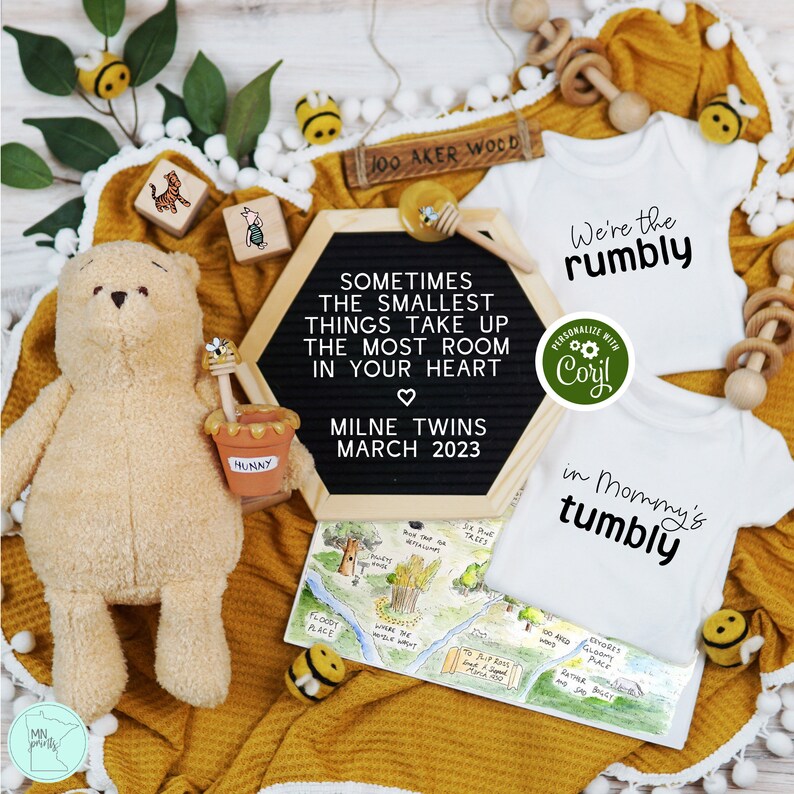 May include: A Winnie the Pooh themed baby announcement with two white onesies. One onesie says "We're the rumbly" and the other says "in Mommy's tumbly". The onesies are surrounded by a yellow blanket, a wooden sign that says "100 Aker Wood", a stuffed Winnie the Pooh bear, and a chalkboard sign that says "Sometimes the smallest things take up the most room in your heart. Milne Twins March 2023".