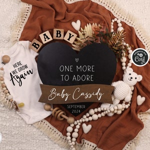 Op de afbeelding: Een witte romper met de tekst "Here we grow again" in zwarte letters. Een zwart hartvormig krijtbord met de tekst "One more to adore" en een houten banner met de tekst "Baby Cassidy September 2024". Houten blokken die "BABY" spellen en een witte gehaakte teddybeer.