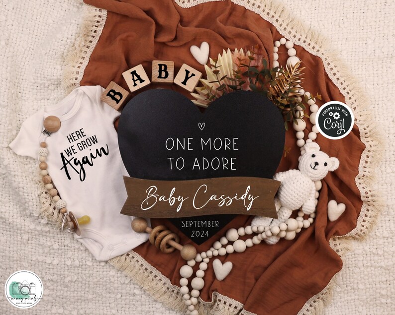 Op de afbeelding: Een witte romper met de tekst "Here we grow again" in zwarte letters. Een zwart hartvormig krijtbord met de tekst "One more to adore" en een houten banner met de tekst "Baby Cassidy September 2024". Houten blokken die "BABY" spellen en een witte gehaakte teddybeer.
