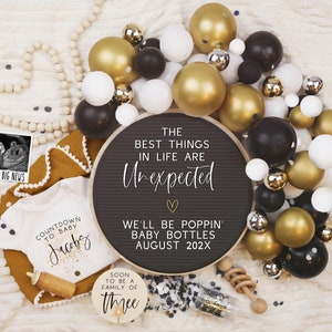 Pode incluir: Um quadro de letras preto com o texto "The best things in life are unexpected" e "We'll be poppin' baby bottles August 202X". O quadro &eacute; rodeado por bal&otilde;es dourados e pretos, uma guirlanda de contas de madeira brancas e um macac&atilde;o branco com o texto "Countdown to baby Jacoby".
