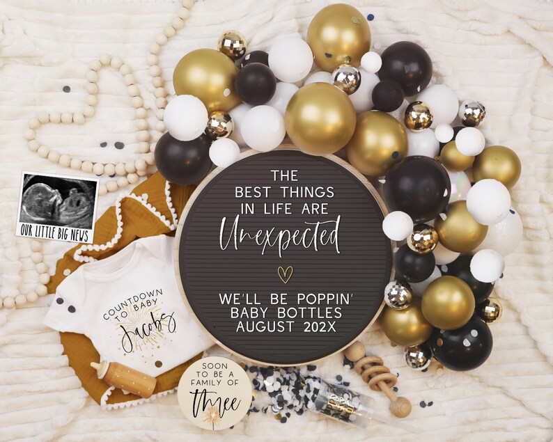 Pode incluir: Um quadro de letras preto com o texto "The best things in life are unexpected" e "We'll be poppin' baby bottles August 202X". O quadro &eacute; rodeado por bal&otilde;es dourados e pretos, uma guirlanda de contas de madeira brancas e um macac&atilde;o branco com o texto "Countdown to baby Jacoby".