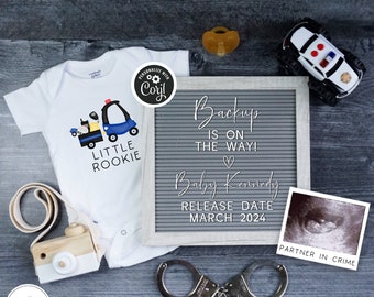 Pode incluir: Um macacão branco com um gráfico de carro de polícia azul e amarelo e o texto "Little Rookie". Um quadro de letras com o texto "Backup is on the way!" e "Baby Kennedy Release Date March 2024". Também estão incluídos na imagem um carro de polícia de brinquedo, uma chupeta e algemas.