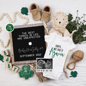 May include: A white onesie with the text "Oops... We Did It Again" in green lettering. The onesie is surrounded by baby items including a wooden block spelling "baby", a rattle, a stuffed lion, and a shamrock-shaped sign that reads "Lucky and Blessed".