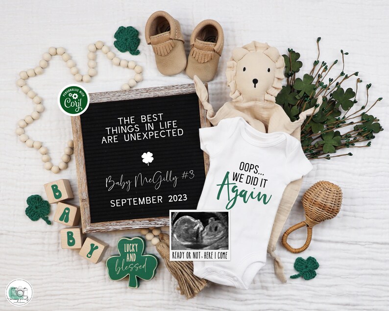 May include: A white onesie with the text "Oops... We Did It Again" in green lettering. The onesie is surrounded by baby items including a wooden block spelling "baby", a rattle, a stuffed lion, and a shamrock-shaped sign that reads "Lucky and Blessed".