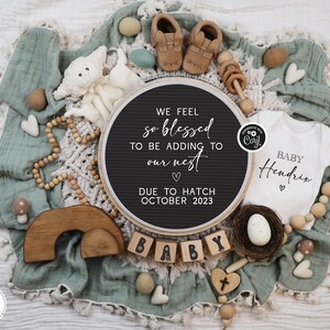 May include: A black felt letter board with the text "WE FEEL so blessed TO BE ADDING TO our nest ♡ DUE TO HATCH OCTOBER 2023". The board is surrounded by a variety of baby items, including a white onesie with the text "BABY Hendrix ♡", a wooden rainbow, a wooden rattle, and a nest with a white egg.