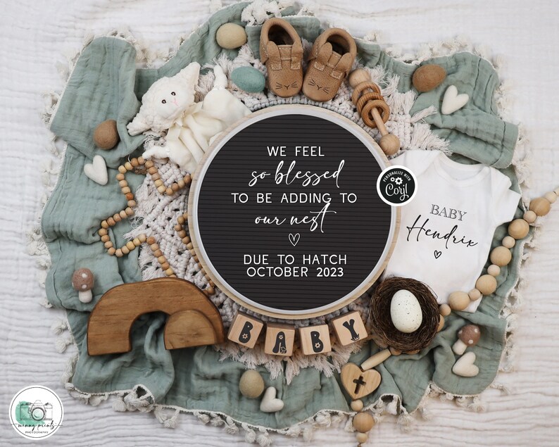 May include: A black felt letter board with the text "WE FEEL so blessed TO BE ADDING TO our nest ♡ DUE TO HATCH OCTOBER 2023". The board is surrounded by a variety of baby items, including a white onesie with the text "BABY Hendrix ♡", a wooden rainbow, a wooden rattle, and a nest with a white egg.