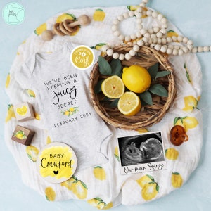 Op de afbeelding: Een grijze romper met de tekst "We've been keeping a juicy secret February 2023". Een houten cirkel met de tekst "Baby Crawford" en een hart. Een houten blok met een citroenontwerp. Een mand met citroenen. Een ingelijst echografiebeeld met de tekst "Our main squeeze".