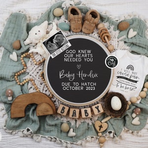 May include: A black letter board with the text "God knew our hearts needed you" and "Baby Hendrix due to hatch October 2023". The board is surrounded by a variety of baby items, including a white onesie with the text "I will not cause pain without allowing something new to be born", a wooden rainbow, a wooden cross, and a small stuffed animal.