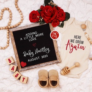 K&ouml;nnte beinhalten: Ein schwarzes Buchstabenbrett mit dem Text "Adding a little more love" und "Baby Hartley August 2023". Ein rotes Herz befindet sich neben dem Text. Ein wei&szlig;er Strampler mit dem Text "Here we grow again" in Rot. Eine Holzrassel und ein Paar braune Babyschuhe sind ebenfalls im Bild.