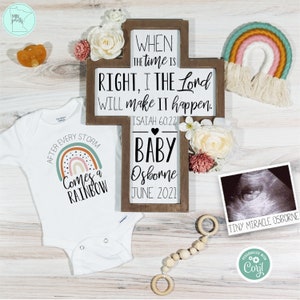 May include: A baby announcement with a white babygrow featuring a rainbow graphic and the text "After Every Storm Comes a Rainbow." A wooden cross-shaped sign displays the text "When the time is right, I the Lord will make it happen." A sonogram is also included.
