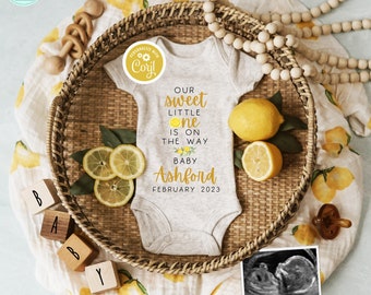 May include: A white onesie with the text "Our sweet little one is on the way Baby Ashford February 2023" on it. The onesie is surrounded by lemons and a wooden tray. There is a small ultrasound image with the text "Our main squeeze" below it.