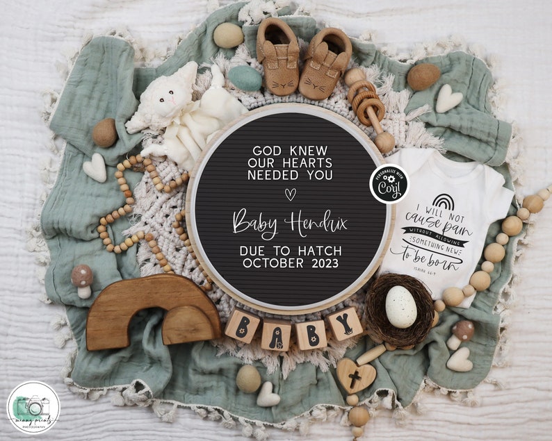 May include: A black letter board with the text "God knew our hearts needed you" and "Baby Hendrix due to hatch October 2023". A white onesie with the text "I will not cause pain without allowing something new to be born" is next to the letter board. The onesie is surrounded by wooden toys, including a rainbow, a nest with an egg, and a wooden cross.
