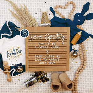 May include: A wooden sign with the text "We're Expecting 2023 to be a great year! Baby Clark #2 Due to arrive July 202X". The sign is surrounded by a white baby onesie with the text "Little Brother", a blue baby blanket, a wooden rattle, a wooden bunny, and a wooden bead garland.