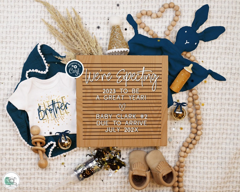 May include: A wooden sign with the text "We're Expecting 2023 to be a great year! Baby Clark #2 Due to arrive July 202X". The sign is surrounded by a white baby onesie with the text "Little Brother", a blue baby blanket, a wooden rattle, a wooden bunny, and a wooden bead garland.