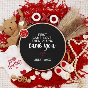 May include: A black chalkboard sign with the text "First came love, then along came you" and the date "July 20XX". The sign is surrounded by red fabric, a teddy bear, a baby onesie, wooden beads, and other Valentine's Day decorations.