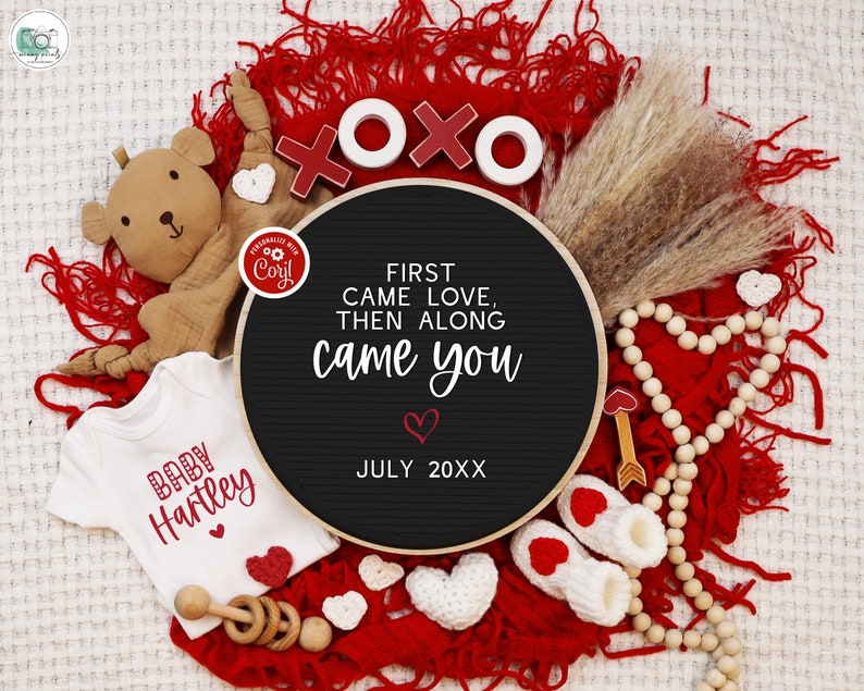 May include: A black chalkboard sign with the text "First came love, then along came you" and the date "July 20XX". The sign is surrounded by red fabric, a teddy bear, a baby onesie, wooden beads, and other Valentine's Day decorations.