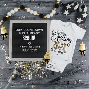 May include: A white onesie with gold lettering that reads "We're Expecting 202X to be great!!" with two gold hearts. A black letter board with white letters reads "Our countdown has already begun! Baby Bennet July 202X". There are gold and silver confetti, black and white baby shoes, and two gold bottle-shaped ornaments with black bows.
