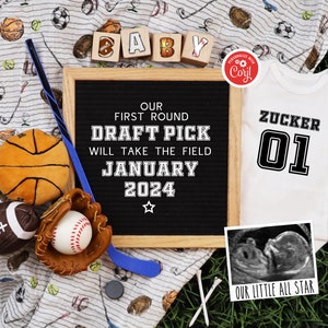 May include: A black letter board with the text "Our First Round Draft Pick Will Take The Field January 2024" surrounded by sports-themed baby items, including a white onesie with the text "Zucker 01" and a framed ultrasound image with the text "Our Little All Star".