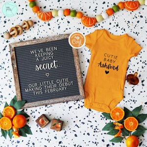 Op de afbeelding: Een letterbord met de tekst "We've been keeping a juicy secret... Our little cutie making their debut this February."  Naast het letterbord ligt een klein oranje rompertje met de tekst "Cutie Baby Ashford."  Het rompertje ligt op een witte achtergrond met sinaasappelschijfjes en groene bladeren.