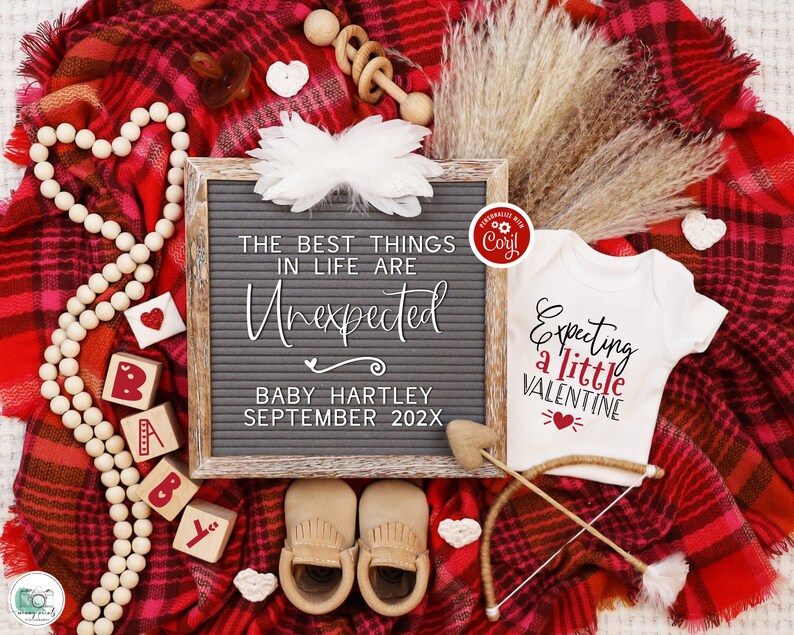 K&ouml;nnte beinhalten: Eine rot-wei&szlig; karierte Decke mit einer Buchstabentafel, auf der steht "Die besten Dinge im Leben sind unerwartet. Baby Hartley September 202X". Es gibt einen wei&szlig;en Strampler mit dem Text "Erwarten ein kleines Valentinstag" und einen Holzpfeil mit einem Herzen darauf. Au&szlig;erdem gibt es Holzkl&ouml;tze, die "Baby" buchstabieren, eine Holzperlenkette und eine Holzrassel.