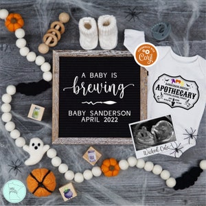 May include: A black letter board with the text "A baby is brewing, Baby Sanderson, April 2022". A white onesie with the text "Sanderson Sisters Apothecary" and a black and white ultrasound image are on a gray wood background with a white garland, a black bat, a white ghost, a small orange pumpkin, and a small green cauldron.