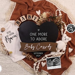 Op de afbeelding: Een zwart hartvormig schoolbord met de tekst "One More To Adore" en "Baby Cassidy" met de datum september 2024. Een witte romper met de tekst "Here We Grow Again" is op de achtergrond. De afbeelding is omringd door houten kralen, een witte teddybeer en een klein echografiebeeld met de tekst "Hello Little Love".