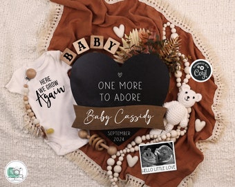 Op de afbeelding: Een zwart hartvormig schoolbord met de tekst "One More To Adore" en "Baby Cassidy" met de datum september 2024. Een witte romper met de tekst "Here We Grow Again" is op de achtergrond. De afbeelding is omringd door houten kralen, een witte teddybeer en een klein echografiebeeld met de tekst "Hello Little Love".