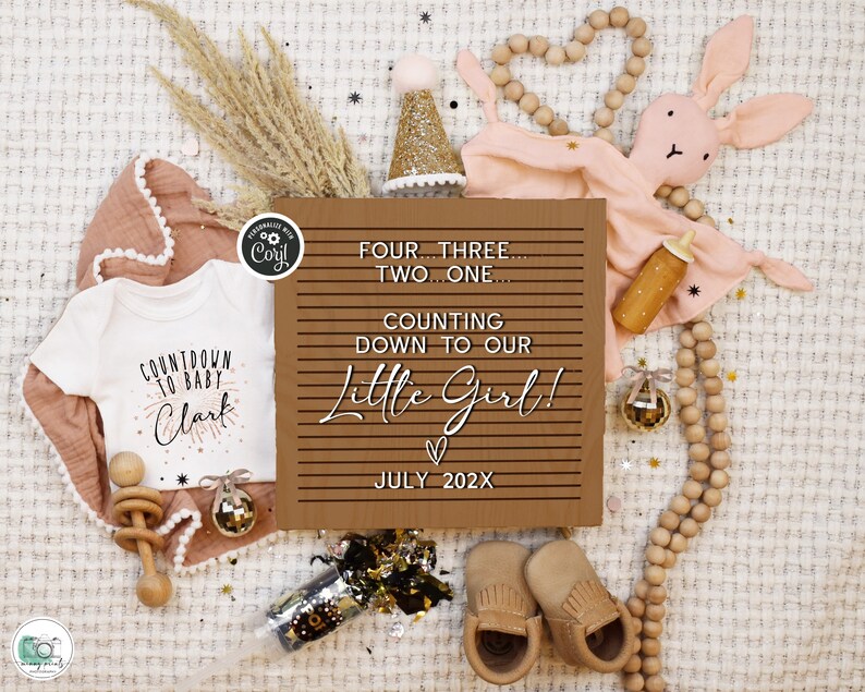 May include: A brown wooden sign with white text that reads "Four...Three...Two...One...Counting down to our Little Girl!  ♡ July 202X".  The sign is surrounded by baby items, including a white onesie with the text "Countdown to Baby Clark", a pink bunny rattle, a wooden rattle, and a gold party hat.
