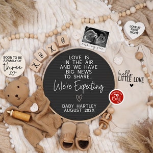 K&ouml;nnte beinhalten: Ein schwarzes Schild aus Schiefer mit dem Text "Love is in the air and we have big news to share... We're Expecting Baby Hartley August 202X". Das Schild ist umgeben von Holzkl&ouml;tzen, einem Teddyb&auml;r, einem Baby-Strampler und einer Babyrassel.