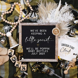May include: A black letter board with the text "We've been keeping a little secret... We'll be poppin' baby bottles July 202X". The board is surrounded by gold and black decorations, including a baby onesie with the text "Countdown to baby Clark", a pair of baby shoes, and a wooden rattle.