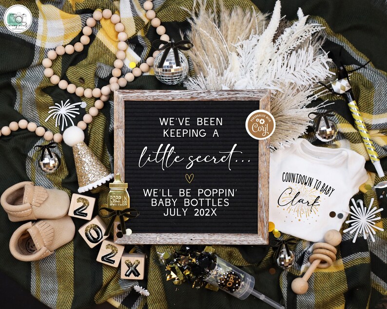 May include: A black letter board with the text "We've been keeping a little secret... We'll be poppin' baby bottles July 202X". The board is surrounded by gold and black decorations, including a baby onesie with the text "Countdown to baby Clark", a pair of baby shoes, and a wooden rattle.