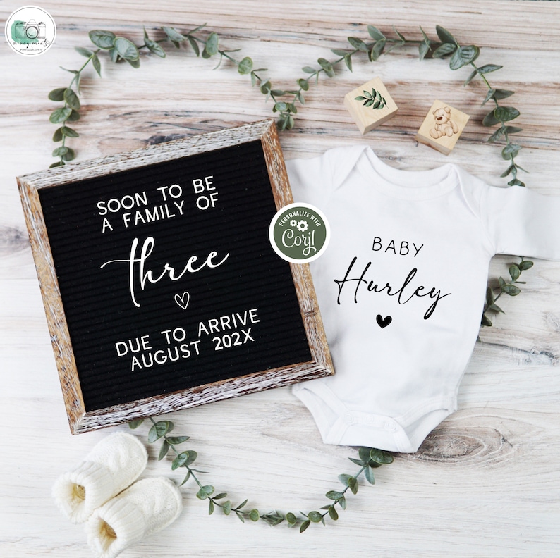 May include: A white onesie with the text "BABY Hurley" in black script with a small black heart below. A black letter board with white letters reads "SOON TO BE A FAMILY OF Three DUE TO ARRIVE AUGUST 202X".