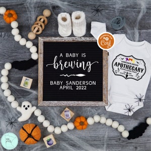 May include: A black letter board with the text "A baby is brewing, Baby Sanderson, April 2022". A white onesie with the text "Sanderson Sisters Apothecary, Potions, Elixirs, Tonics, Est. 1693, Salem, Mass."  A white ghost, a pumpkin, and a cauldron are on a gray surface with white spider webs.