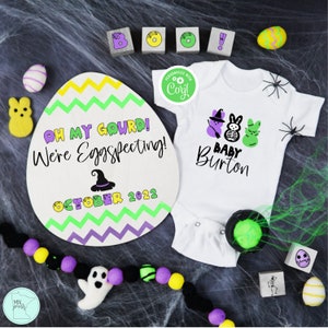 May include: A Halloween-themed pregnancy announcement with a purple, green, and yellow egg-shaped sign that reads "Oh my gourd! We're Eggspecting! October 2022". A white onesie with a purple and green ghost, a skeleton, and a green Peep with the text "Baby Burton" is also included.
