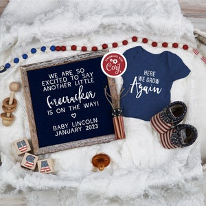 May include: A blue and white letter board with the text "We are so excited to say another little firecracker is on the way! Baby Lincoln January 2023". A blue baby onesie with the text "Here we grow again" is next to the letter board. A red, white, and blue baby rattle, a red, white, and blue beaded necklace, and a red, white, and blue baby shoe are also in the image.